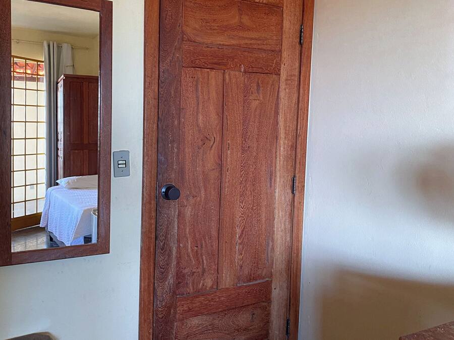 Una puerta de madera con un espejo en la pared, una cama con cubrecama blanco y un pequeño taburete de madera.