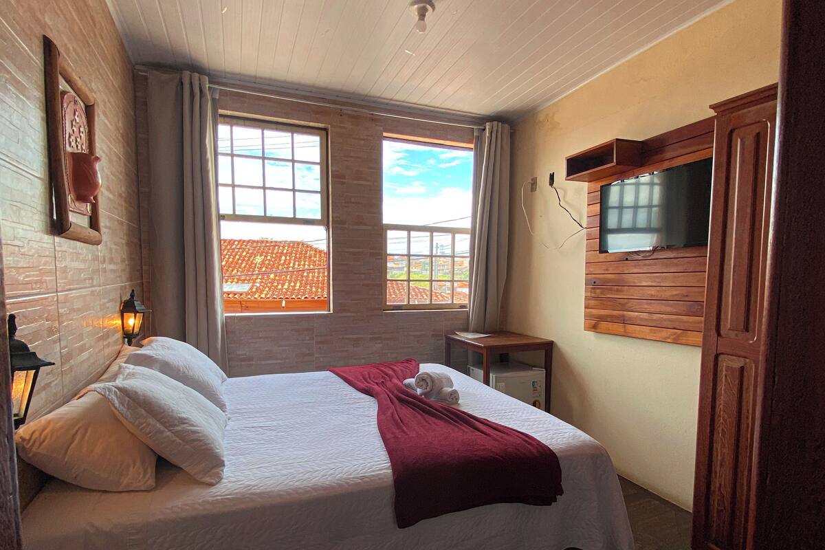 A bedroom with a bed, two windows, a wooden cabinet, and a wall-mounted TV.