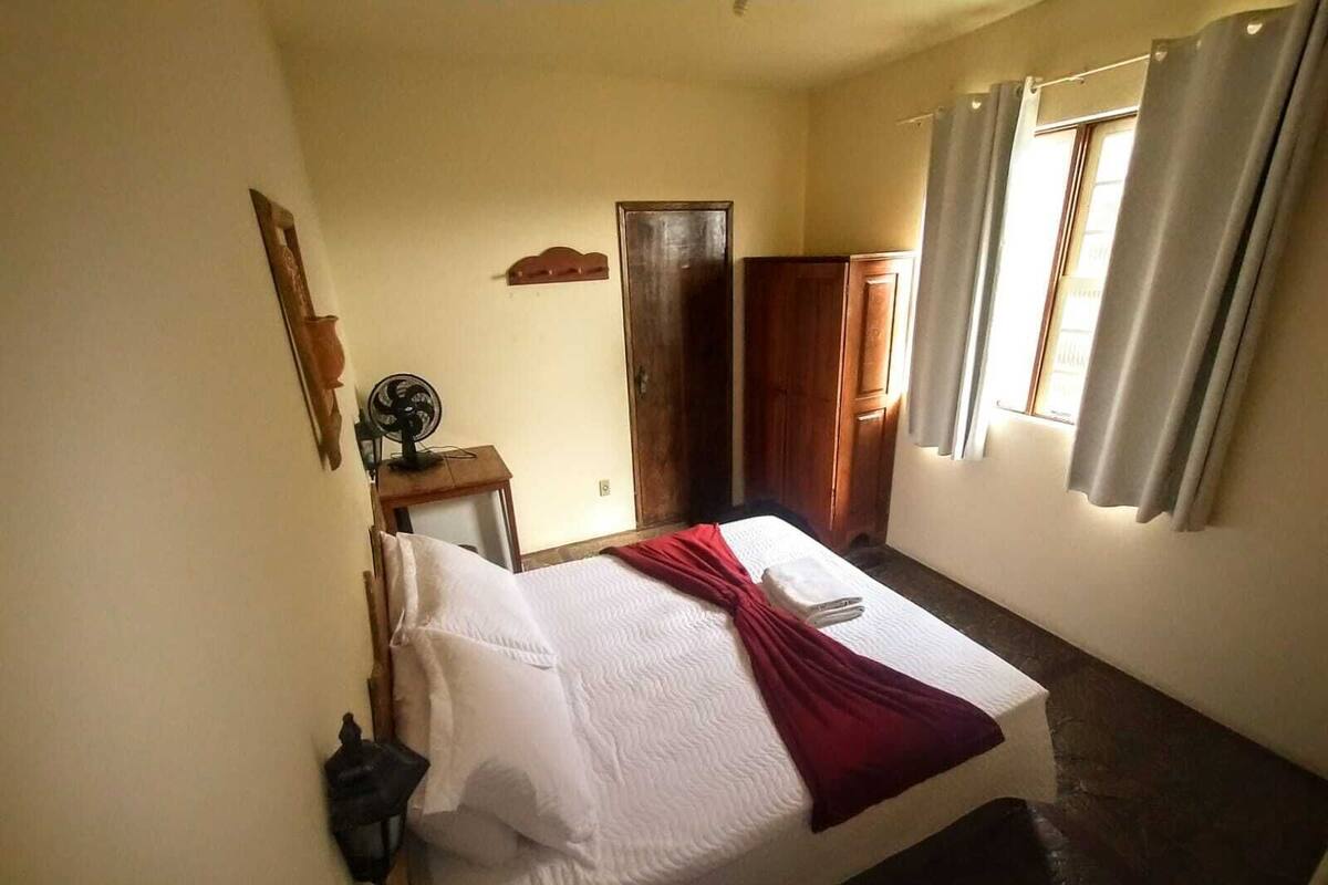 A single bed with white bedding and a red blanket, a bedside table with a fan, a wooden wardrobe, and a window with curtains.