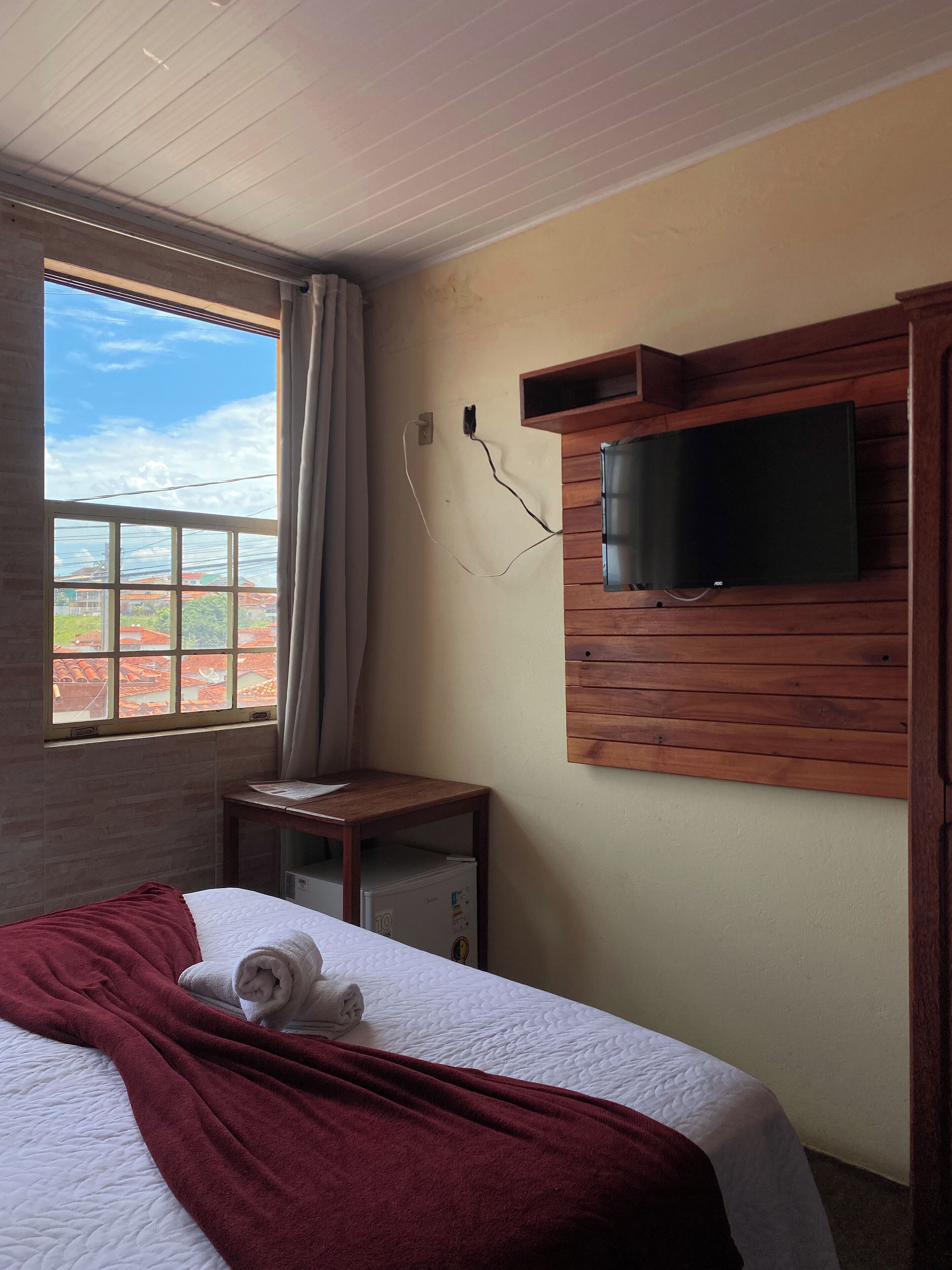 A hotel room with a bed, a TV mounted on a wooden panel, a window with a view of buildings, and a small table with a lamp.