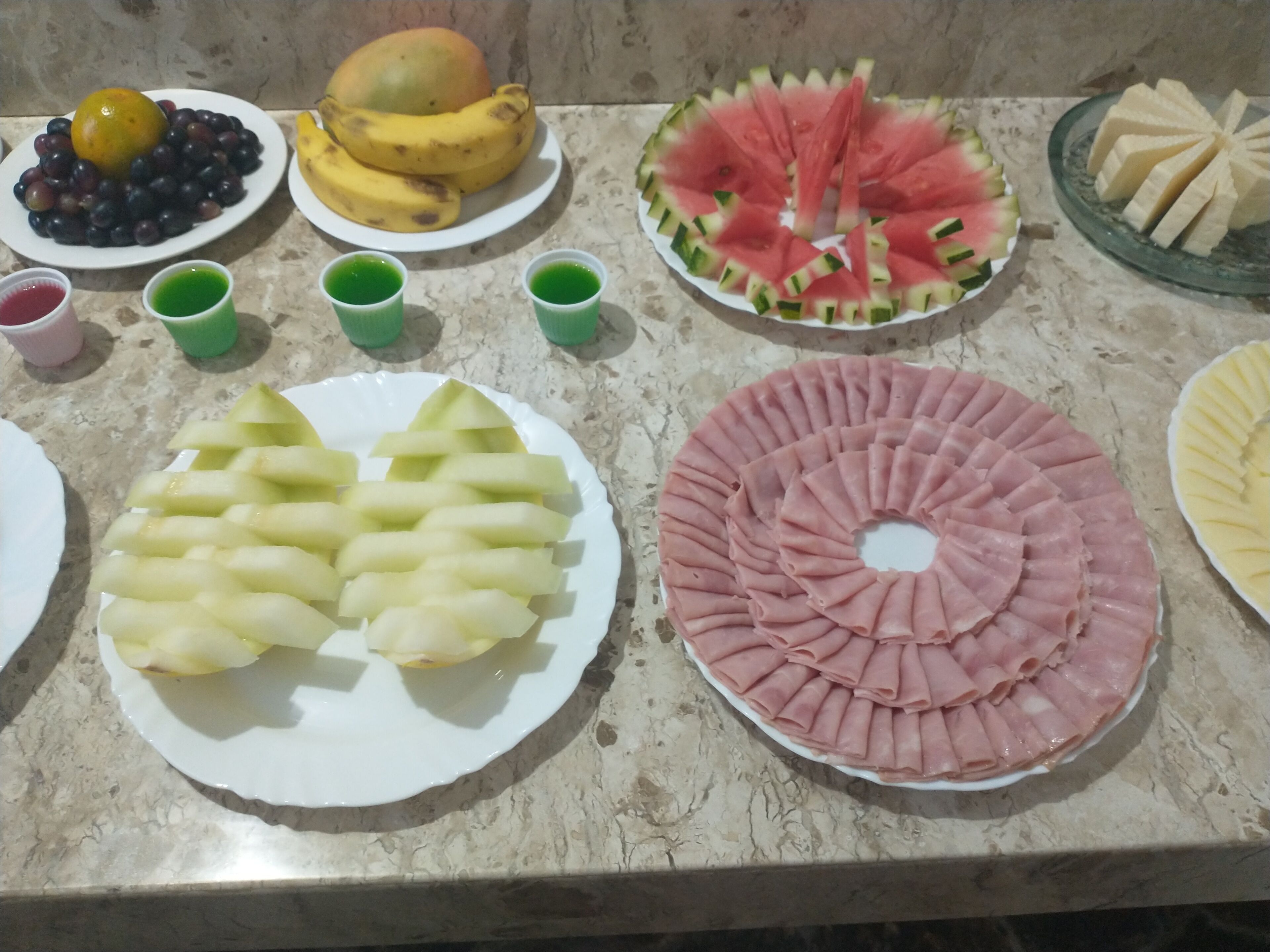 Diversos alimentos dispostos em uma bancada, incluindo pratos com frutas fatiadas, uma melancia e um prato com carne fatiada.
