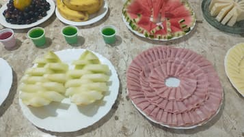 Diversos alimentos dispostos em uma bancada, incluindo pratos com frutas fatiadas, uma melancia e um prato com carne fatiada.