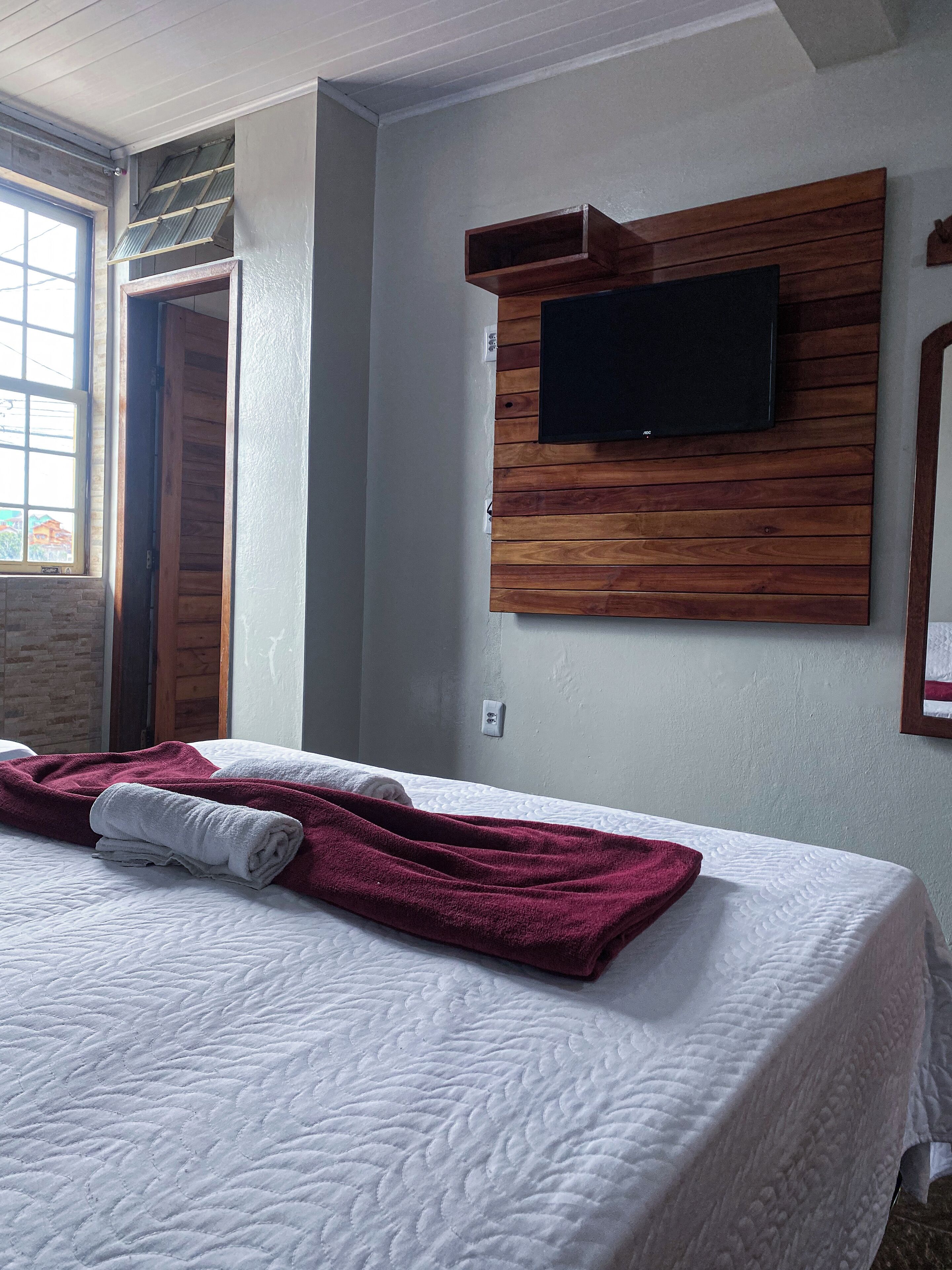 A hotel room with a bed, a TV mounted on a wooden panel, and a window with a view of buildings.