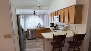 Una cocina con armarios de madera, un lavavajillas de acero inoxidable, un microondas y una cafetera. Hay dos sillas de madera en la isla.