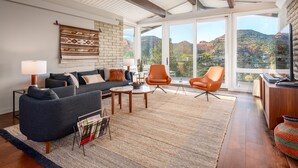 Un salon moderne avec un canapé bleu foncé, des fauteuils orange, une table basse en bois et une grande fenêtre donnant sur un paysage montagneux.