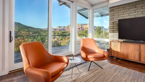 Un salon moderne doté de grandes portes vitrées, de deux fauteuils orange, d’une table basse ronde et d’un téléviseur à écran plat posé sur un meuble en bois.