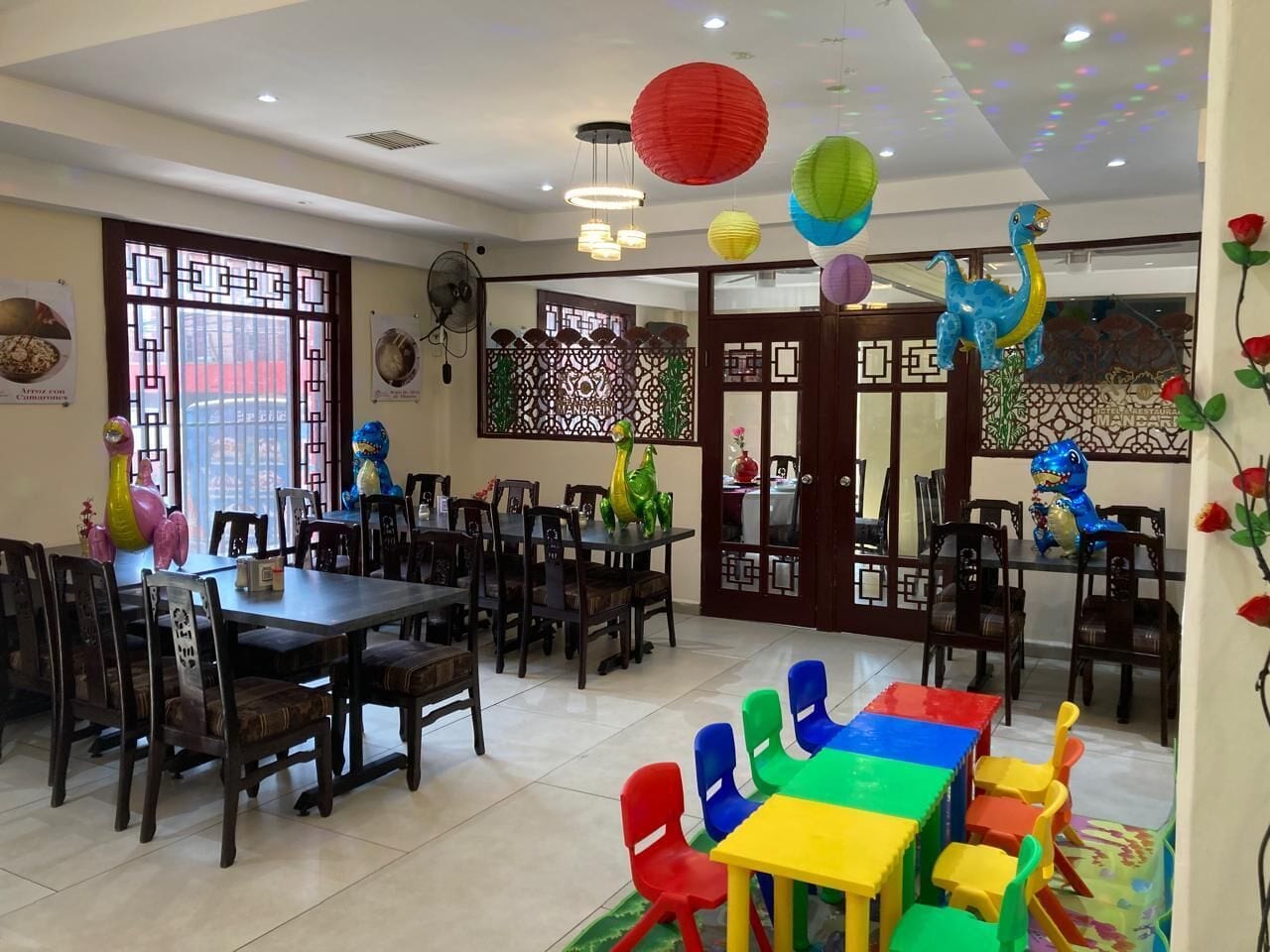 A dining area with wooden tables and chairs, decorative paper lanterns, and a colorful play area with a dinosaur-shaped toy.