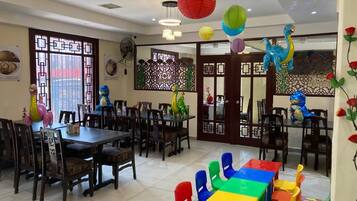 A dining area with wooden tables and chairs, decorative paper lanterns, and a colorful play area with a dinosaur-shaped toy.