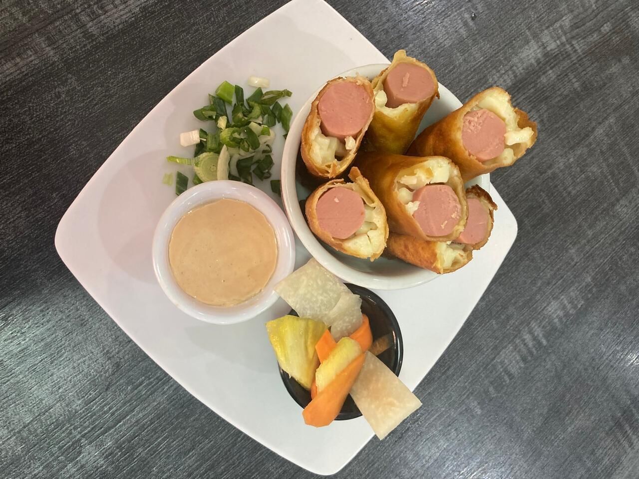 A plate with sausage rolls, a small bowl of sauce, and some vegetables.