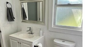 Een badkamer met een wit wastafelmeubel, spiegel en toilet, een vloerkleed en een raam met uitzicht.