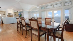 Een ruime eethoek met een grote houten tafel en stoelen, een keuken op de achtergrond en uitzicht op de oceaan door grote ramen.