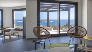 Une chambre moderne avec de grandes fenêtres donnant sur la mer, agrémentée de chaises en osier et d’une table ronde.