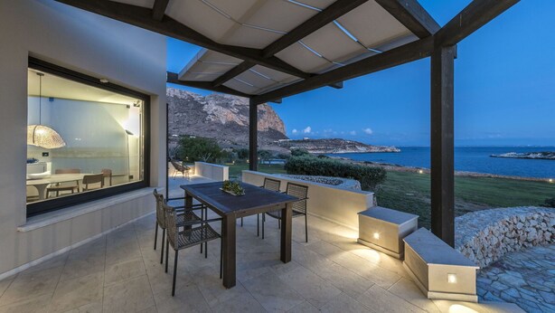 Une terrasse extérieure couverte, dotée d’une table et de chaises, d’un coin salon avec des coussins, et offrant une vue sur la mer et les montagnes.