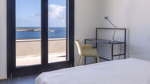 Une chambre d’hôtel avec un lit, un bureau et une chaise, et une vue sur la mer grâce à de grandes fenêtres.