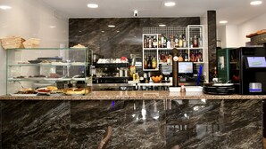 Een moderne hotel lobby met een marmeren receptiebalie, glazen vitrines met gebak, een koffieapparaat en een drankendisplay.