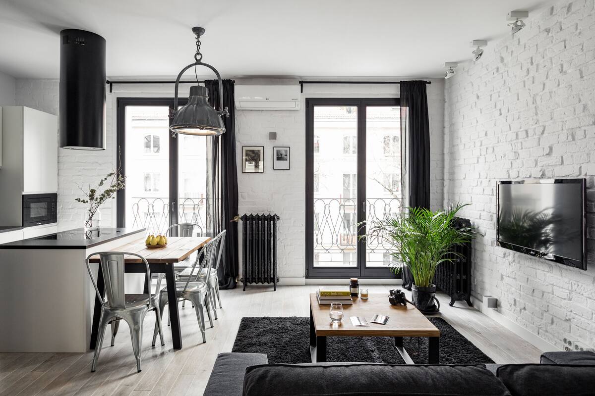 A modern apartment with a kitchen, dining area, and living room. It features a large window, a flat-screen TV, and a potted plant.