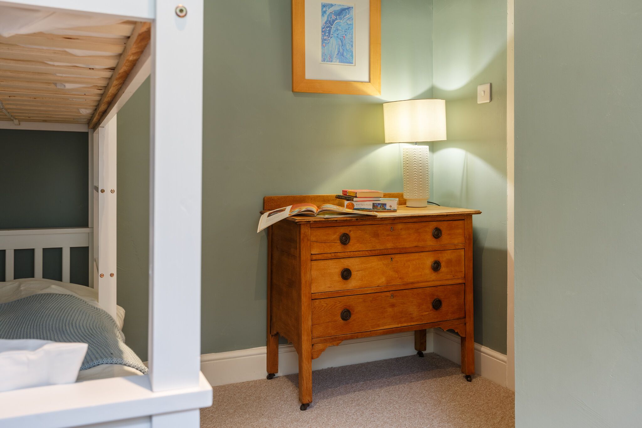 A bedroom with a bunk bed, a wooden dresser with drawers, a bedside table with a lamp, and a framed picture on the wall.