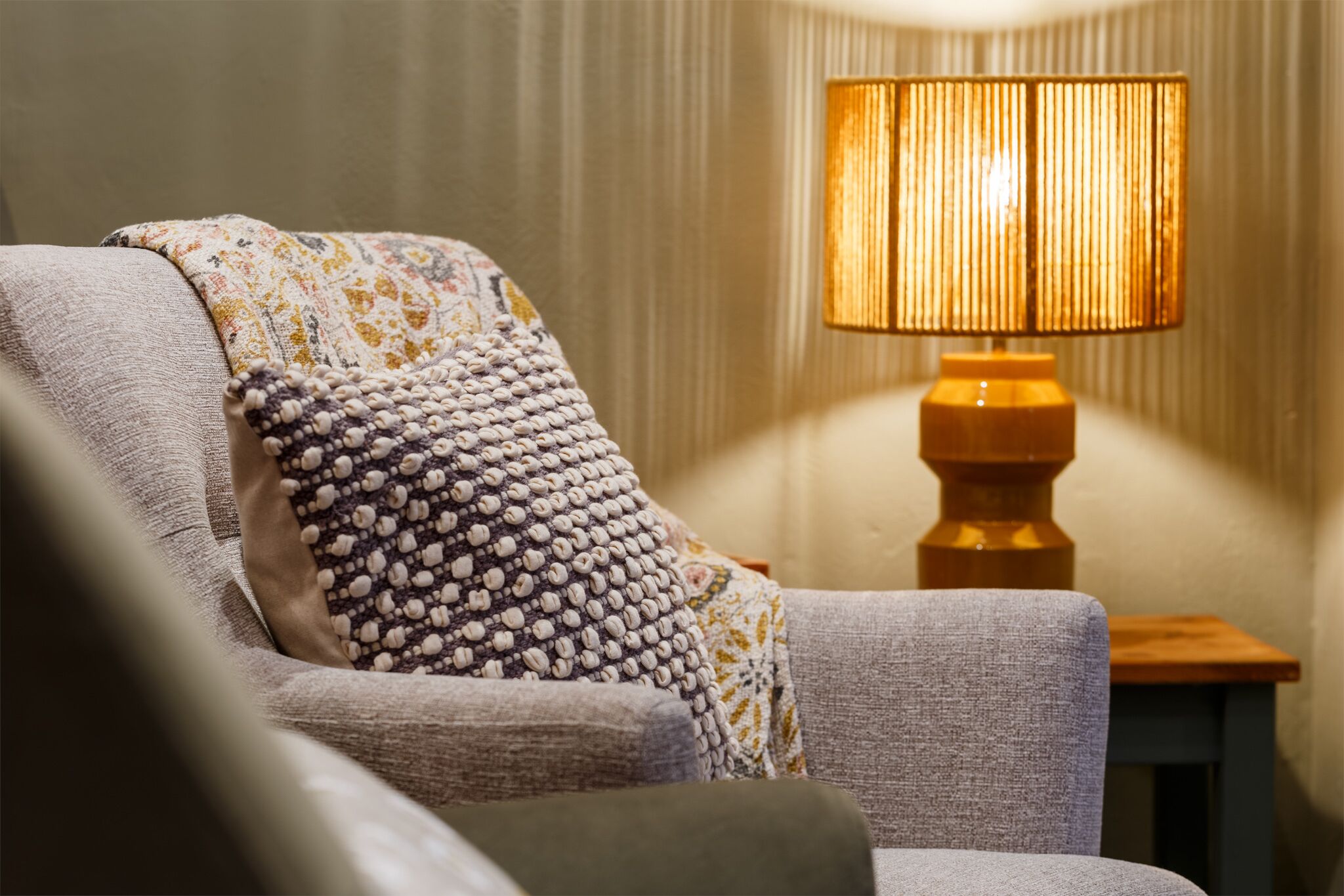 Un fauteuil confortable, agrémenté d’un plaid à motifs et d’une petite table en bois sur laquelle est posée une lampe.