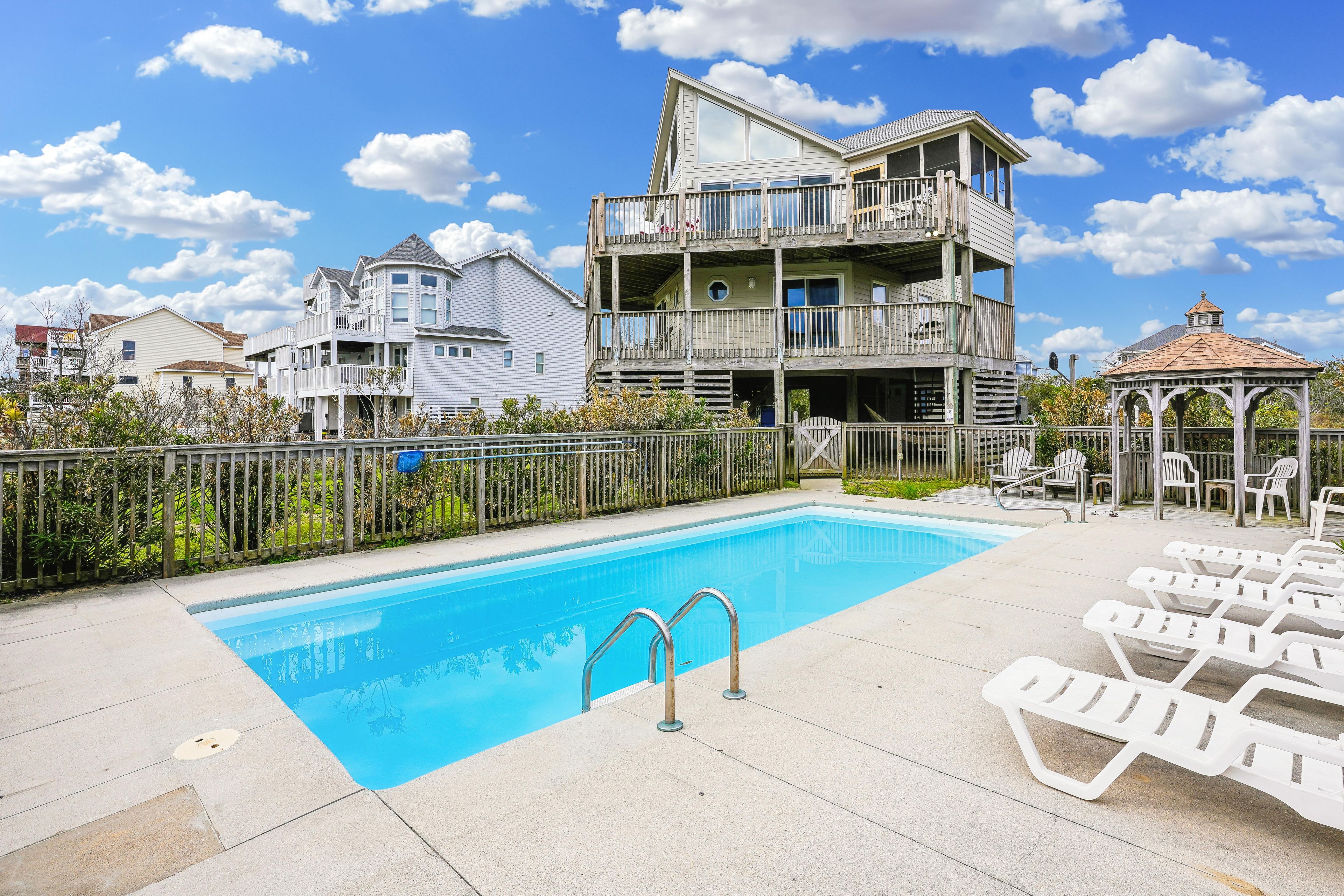 A swimming pool with lounge chairs and a gazebo, surrounded by a wooden fence and a multi-story building with balconies.
