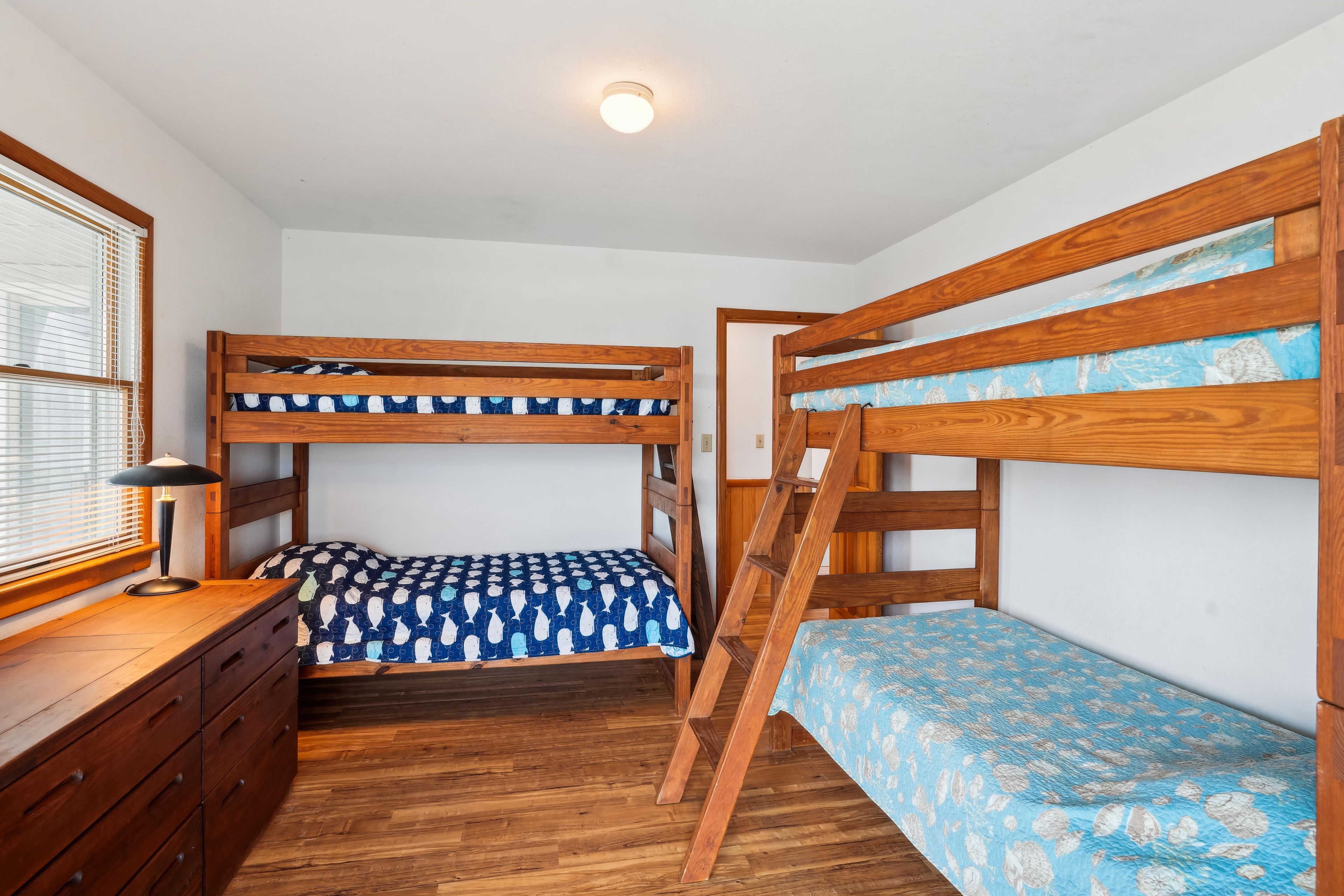 A bunk bed room with a desk and dresser, wooden flooring, and a window with blinds.