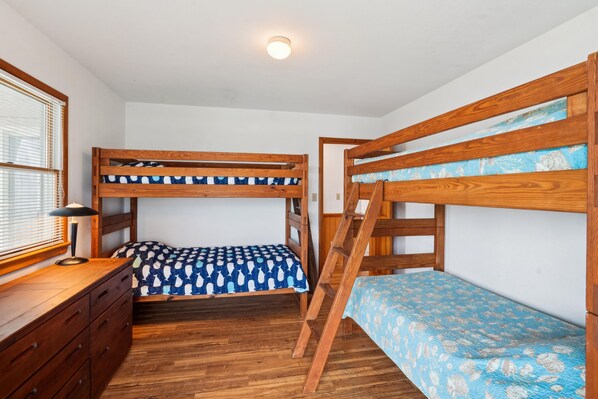 A bunk bed room with a desk and dresser, wooden flooring, and a window with blinds.