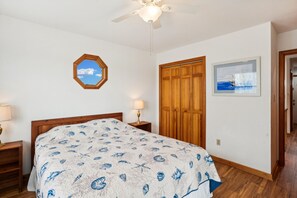 A bedroom with a bed, wooden wardrobe, a ceiling fan, a window with a view, and a framed picture on the wall.