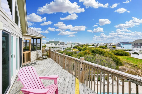 Una terraza de madera con una silla Adirondack rosa, con vista a una zona residencial con casas y un cielo despejado.