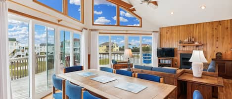 A spacious room with a wooden dining table, blue chairs, a ceiling fan, and large windows offering a view of a residential area.