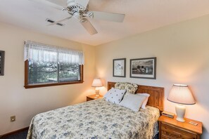 Un dormitorio con una cama, dos lámparas de noche, un ventilador de techo, una ventana con cortinas de encaje y dos cuadros en la pared.