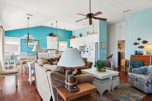 A living room with a ceiling fan, a dining area with a table and chairs, and a kitchen area with cabinets and appliances. - Carolina Hideaway -- Kitty Hawk Home just steps to the beach with Elevator (Kitty Hawk)