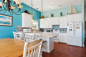 A modern kitchen with white cabinets, a wooden dining table, and a large island. - Carolina Hideaway -- Kitty Hawk Home just steps to the beach with Elevator (Kitty Hawk)