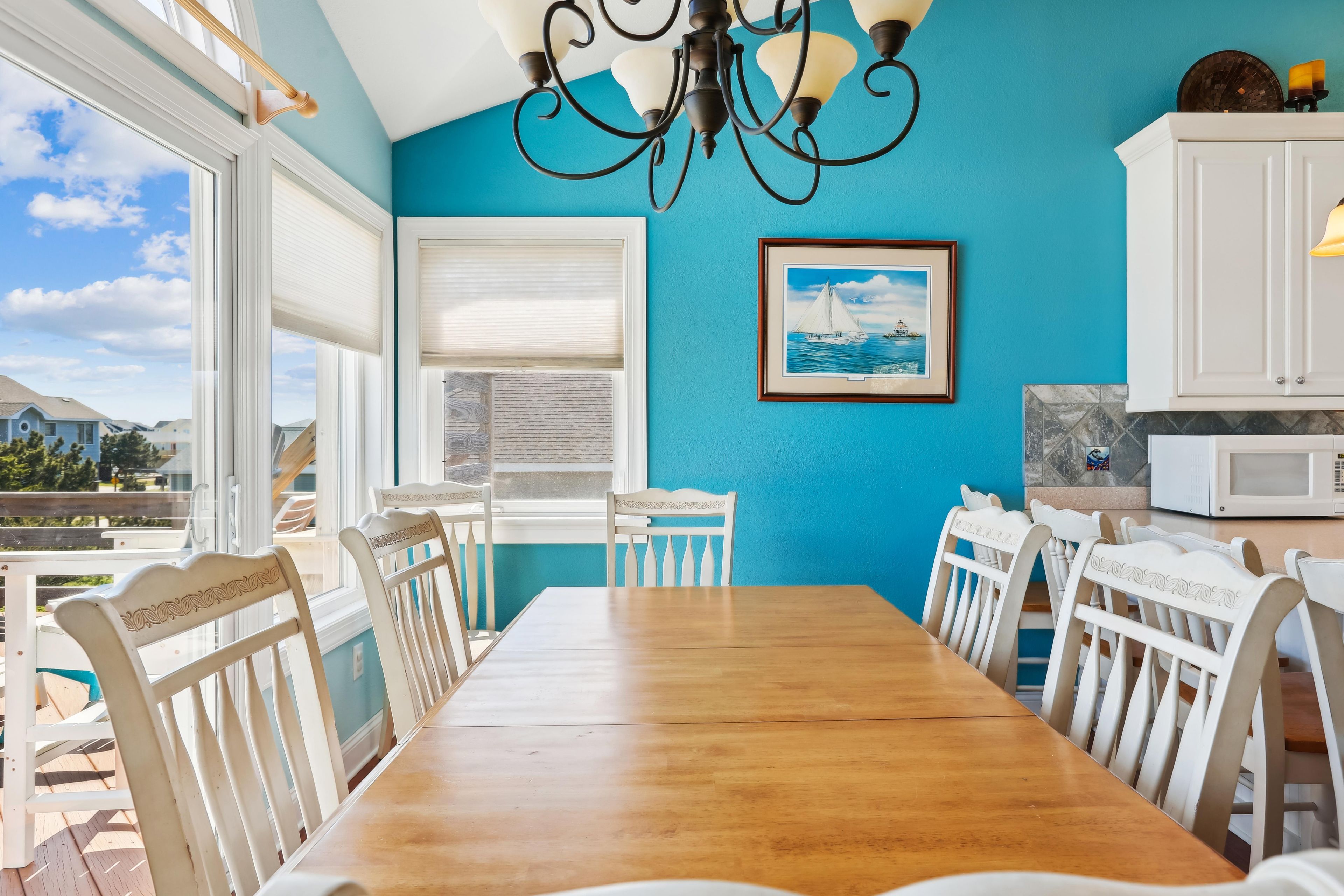 A dining area with a wooden table and white chairs, a blue wall, a framed picture, and a white cabinet with a microwave.