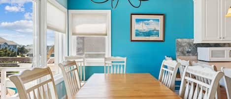 A dining area with a wooden table and white chairs, a blue wall, a framed picture, and a white cabinet with a microwave.