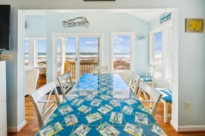 Uma sala de jantar com mesa de vidro exibindo cartões postais com tema náutico, cadeiras brancas e vista para a praia através de grandes janelas.