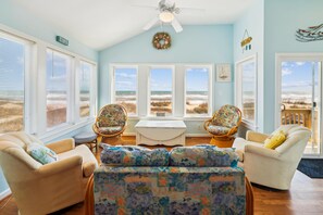 A room with a view of the beach, featuring a ceiling fan, two wicker chairs, a sofa, and a small table. - Lovelady -- Pet Friendly Oceanfront with Hot Tub (Kill Devil Hills)