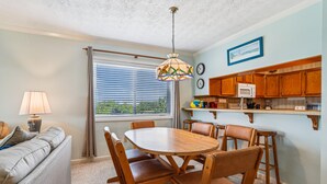 A dining area with a wooden table and chairs, a kitchen with wooden cabinets, and a window with blinds. - Duck Legacy -- 3BR Oceanfront Condo 2nd Floor with Ocean Views (Duck)