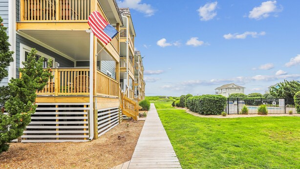 A wooden deck leading to a building with a balcony, a flag, and a well-maintained lawn. - Duck Legacy -- 3BR Oceanfront Condo 2nd Floor with Ocean Views (Duck)