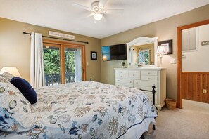 A bedroom with a bed, a dresser, a ceiling fan, a TV, and a window with a view of greenery.