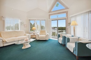 A spacious living room with a high ceiling, large windows, and a view of the outdoors. There are two sofas, a coffee table, and a chair. - Crystal Sands -- Unique 5 BR in Southern Duck with Community Amenities (Duck)