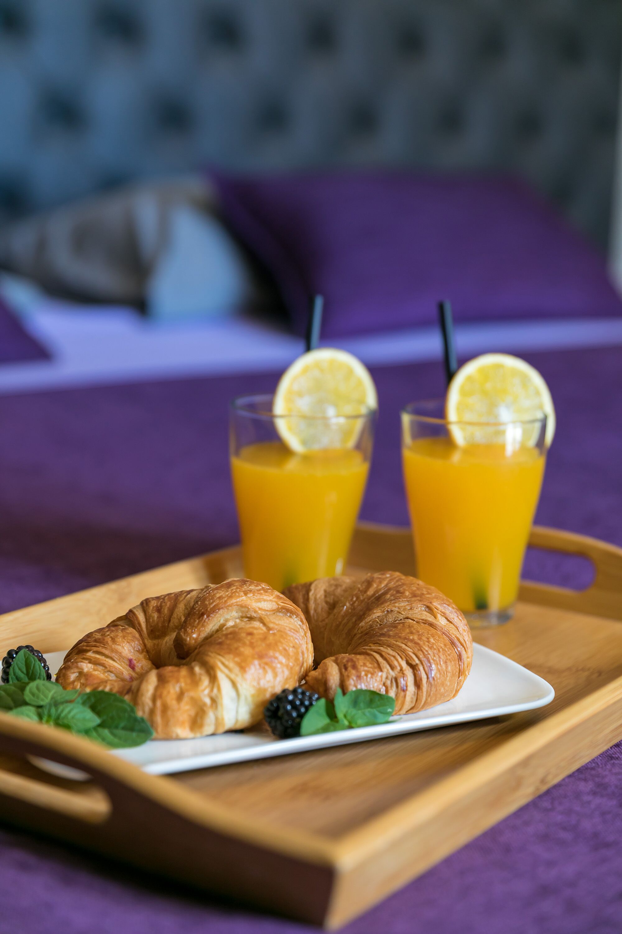Bandeja de café da manhã com croissants e amoras, e dois copos de suco de laranja com rodelas de limão sobre uma mesa de madeira.