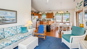 Un salon comprenant un canapé, un fauteuil bleu, une table à manger avec des chaises, un coin cuisine et une vue sur l’extérieur.