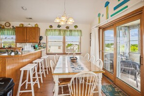 Un espace repas avec une longue table, des chaises blanches et une vue sur l’extérieur, visible à travers une porte vitrée.