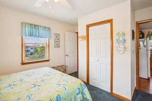 A bedroom with a bed, a window with blinds, a door, and a hallway leading to a kitchen. - Duck Views-- 4BR Oceanside Duck Home with Private Pool (Duck)
