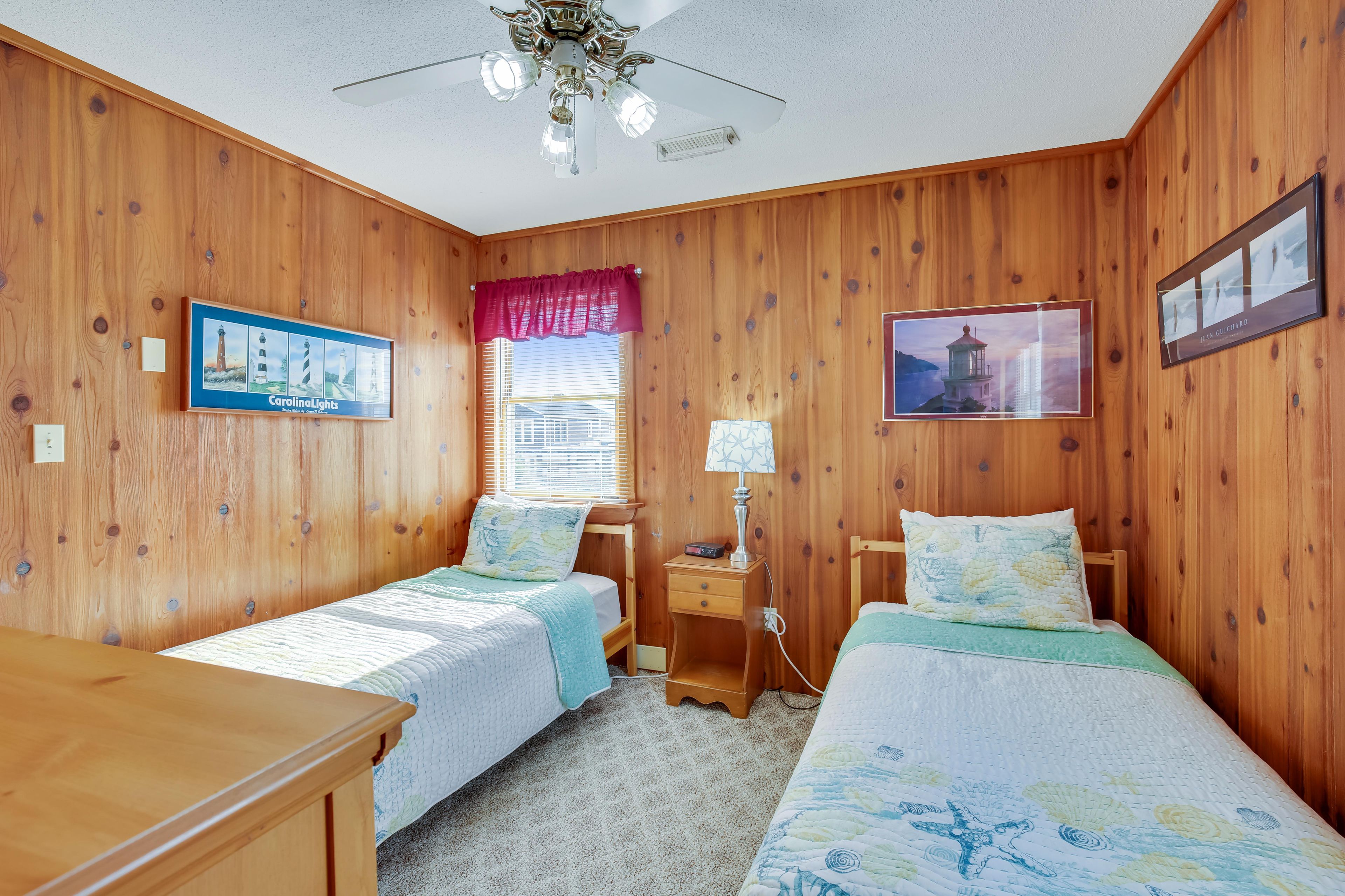 Une pièce aux murs en bois, avec deux lits simples dotés de têtes de lit assorties, un ventilateur de plafond, une fenêtre avec des stores, une table de chevet avec une lampe et deux tableaux encadrés.