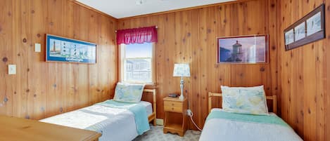 Une pièce aux murs en bois, avec deux lits simples dotés de têtes de lit assorties, un ventilateur de plafond, une fenêtre avec des stores, une table de chevet avec une lampe et deux tableaux encadrés.