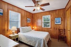 Un dormitorio con paneles de madera, una cama con cubrecama blanco, una mesita de noche con lámpara, un ventilador de techo y dos ventanas con persianas.