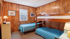 A room with wooden walls, a bunk bed, two bedside tables with lamps, and two framed pictures on the wall. - Sunseeker -- Beat the traffic with a Friday arrival/3 BR Cottage in Kitty Hawk (Kitty Hawk)