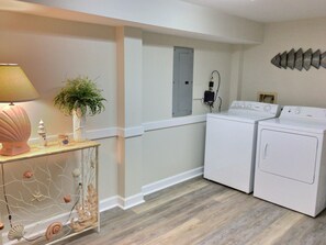 Une buanderie équipée de deux machines à laver blanches, d’une console en bois surmontée d’une lampe et agrémentée d’objets décoratifs, ainsi que d’un tableau électrique fixé au mur.
