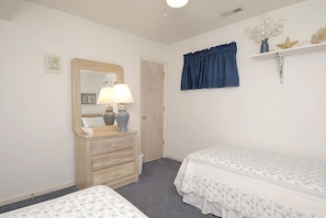 A bedroom with a wooden dresser, two beds with floral bedding, a mirror, a blue curtain, and a shelf with decorative items. - School Bored --Affordable Cottage just 2 blocks to the ocean (Kill Devil Hills)
