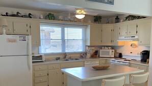 Eine Küche mit weißen Schränken, einem Kühlschrank, einer Mikrowelle, einem Toaster und einer Spüle. An der Kochinsel stehen zwei Stühle und es gibt ein Fenster mit Jalousien.